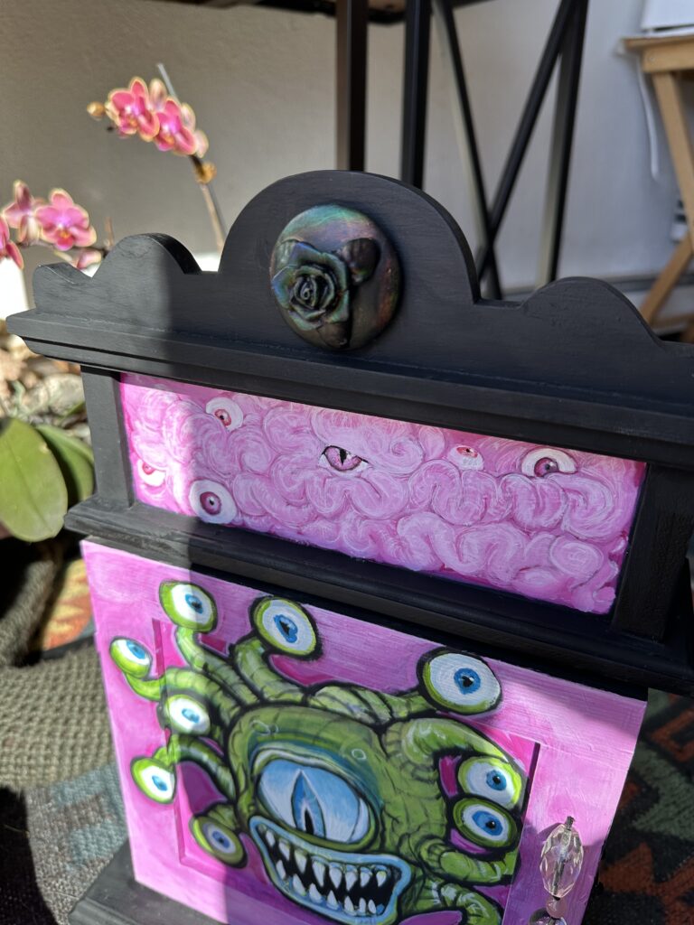 a pink and black cabinet with a beholder on it stands in front of some mini dendrobium orchids.
There is a black rose medallion that appears to have a rainbow effect on it.