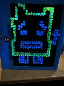 A blue lite-brite toy stands on a wooden surface. On it, in blue and green pegs, is a pixel outline of Ignignokt, a Mooninite from Aqua Teen Hunger Force.
The features are in blue pegs, and the outline is done in green. He is a blocky character, and is also flipping the bird.