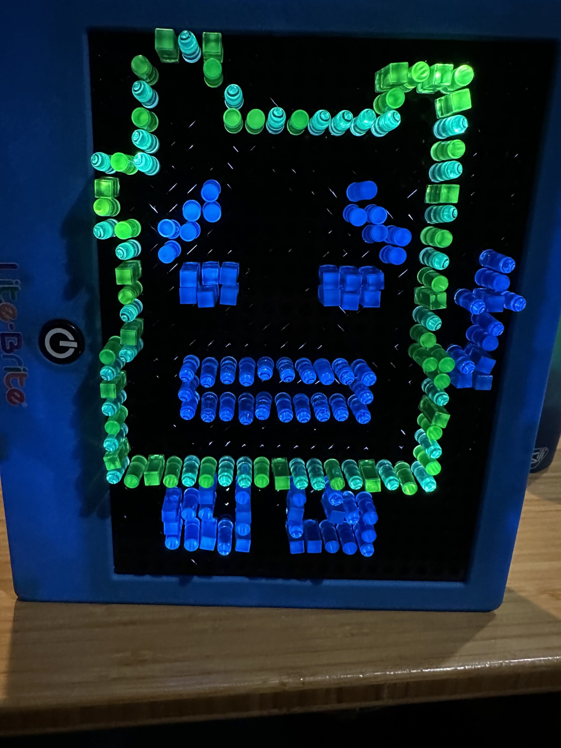 A blue lite-brite toy stands on a wooden surface. On it, in blue and green pegs, is a pixel outline of Ignignokt, a Mooninite from Aqua Teen Hunger Force. The features are in blue pegs, and the outline is done in green. He is a blocky character, and is also flipping the bird.